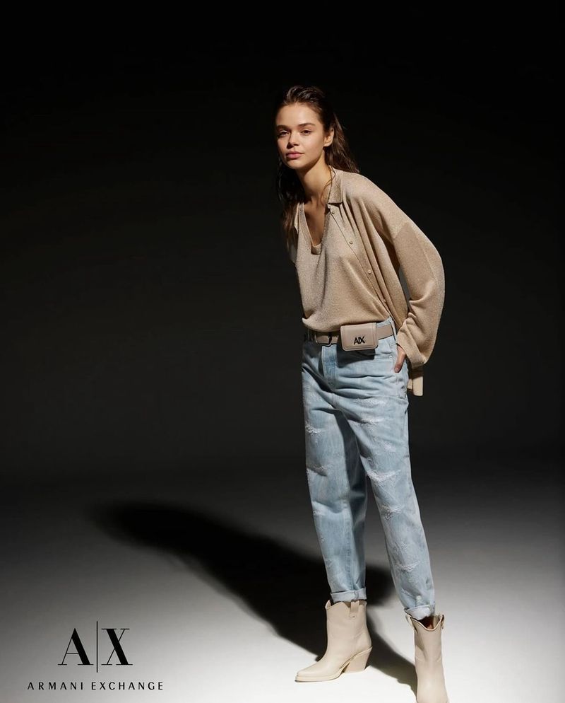 Full-length studio shot of a young adult woman wearing a beige top, light blue jeans, and light-colored boots, standing in a dark studio.