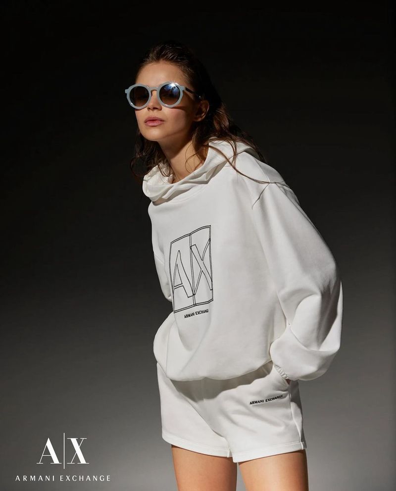 A model wearing a white Armani Exchange hoodie and shorts poses in a studio with sunglasses. Fashion and studio lighting.