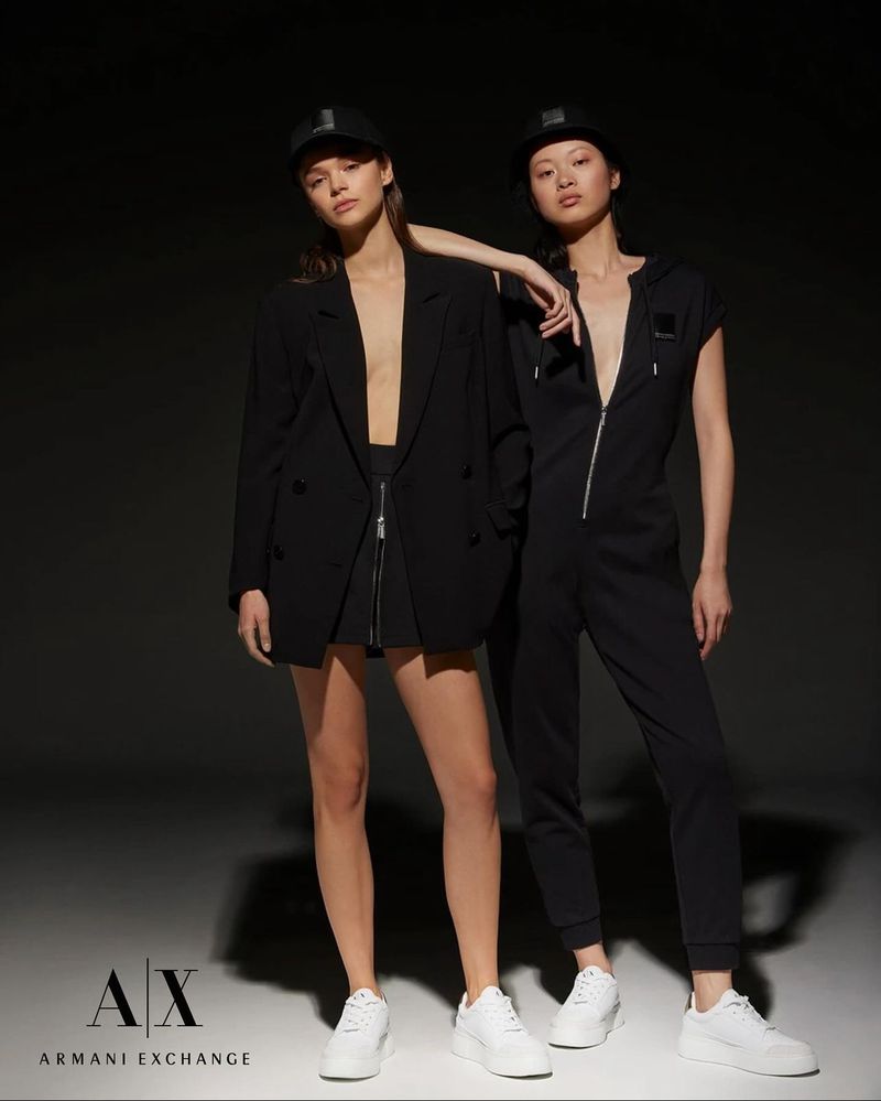 Two women pose in black jumpsuits and matching hats. Fashion-forward attire, white sneakers, studio lighting, neutral backdrop.