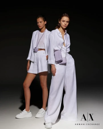 Two fashion models pose indoors wearing white linen outfits with light purple crossbody bags and white sneakers against a dark backdrop.