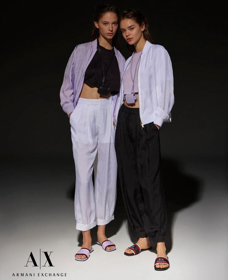 Two fashion models pose in light jackets and pants; one wears lilac, the other black, in a minimalist studio fashion shot.