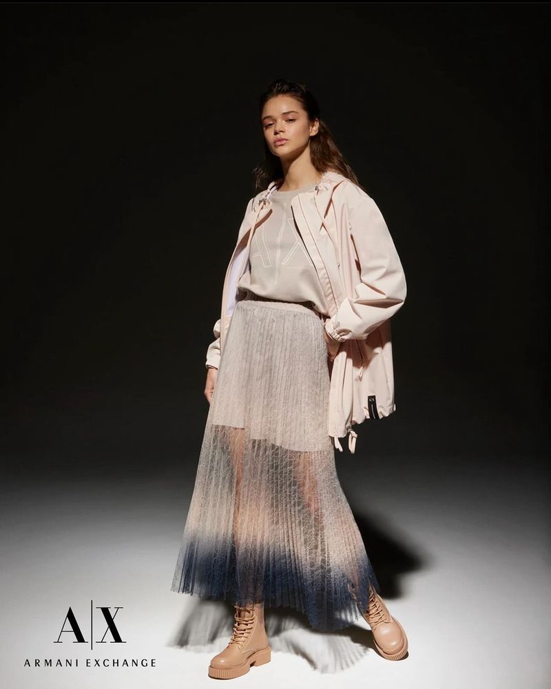 Fashionable brunette woman in a long pleated ombre skirt and jacket posing in a studio with a dark background.