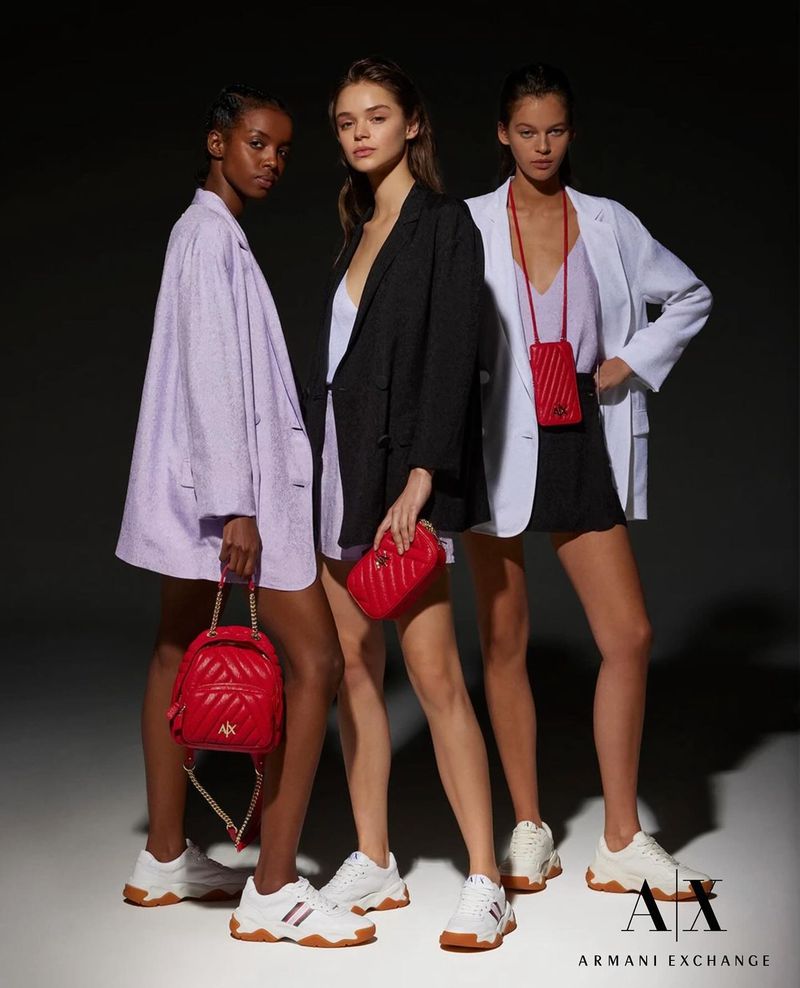 Three fashion models pose wearing blazers, skirts, sneakers, and carrying handbags by Armani Exchange in a studio shot.