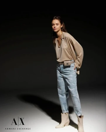 Model posing in light wash denim jeans, a beige knit sweater, and cream ankle boots against a dark studio background.