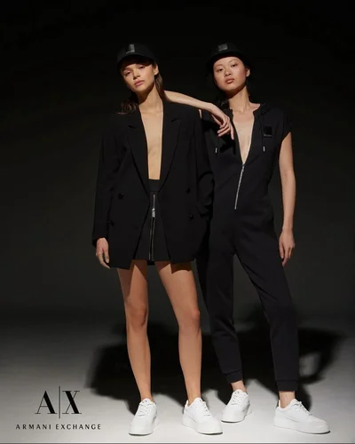 Two female models in black outfits and white sneakers posing against a dark background for Armani Exchange SS24.