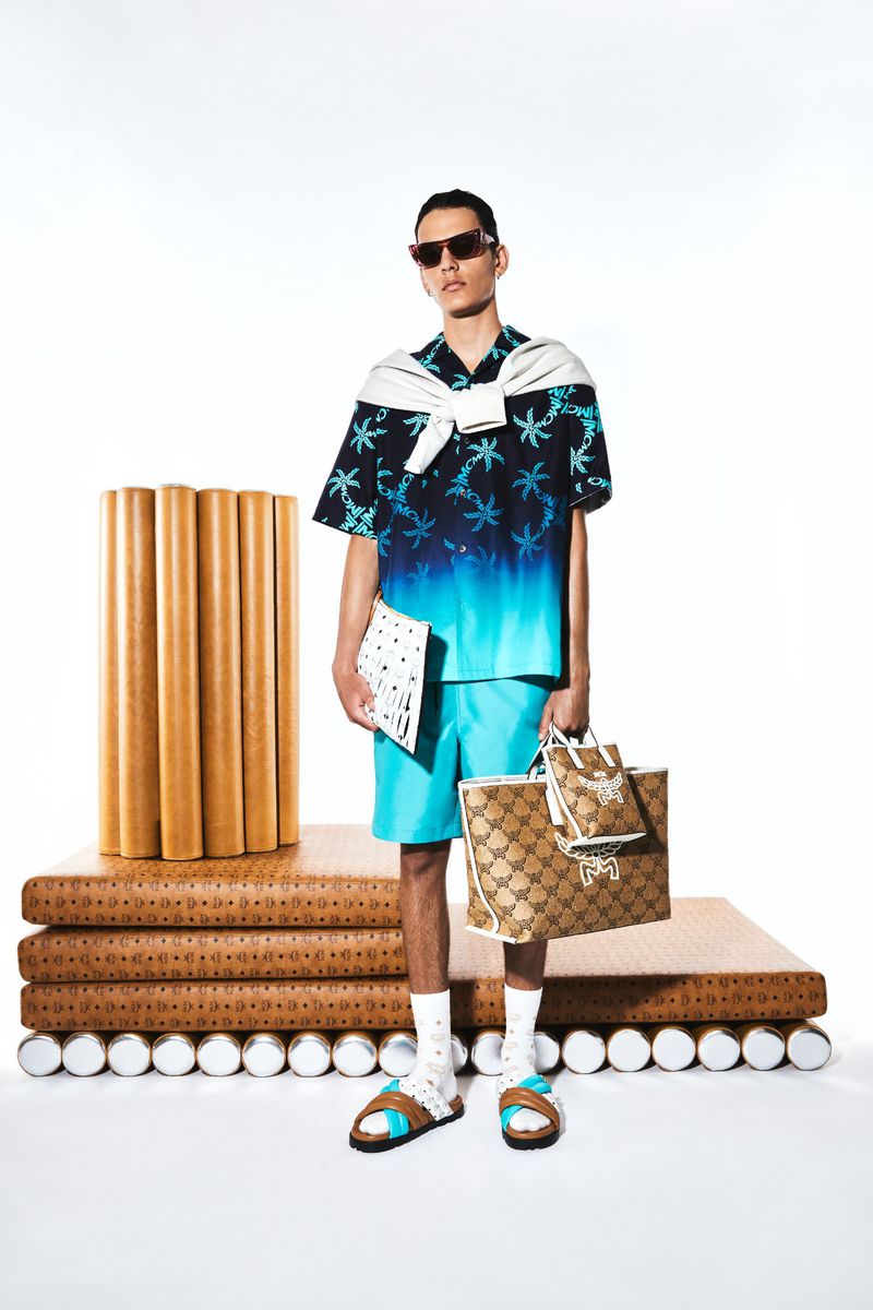 Male fashion model in a blue palm-tree shirt, blue shorts, sunglasses, and slides, carrying shopping bags in a studio setting.
