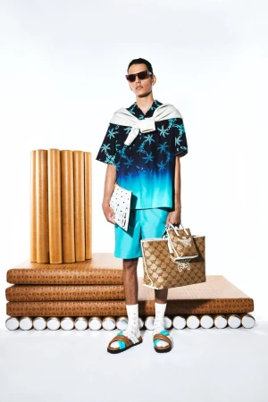 Male fashion model in a blue palm-tree shirt, blue shorts, sunglasses, and slides, carrying shopping bags in a studio setting.