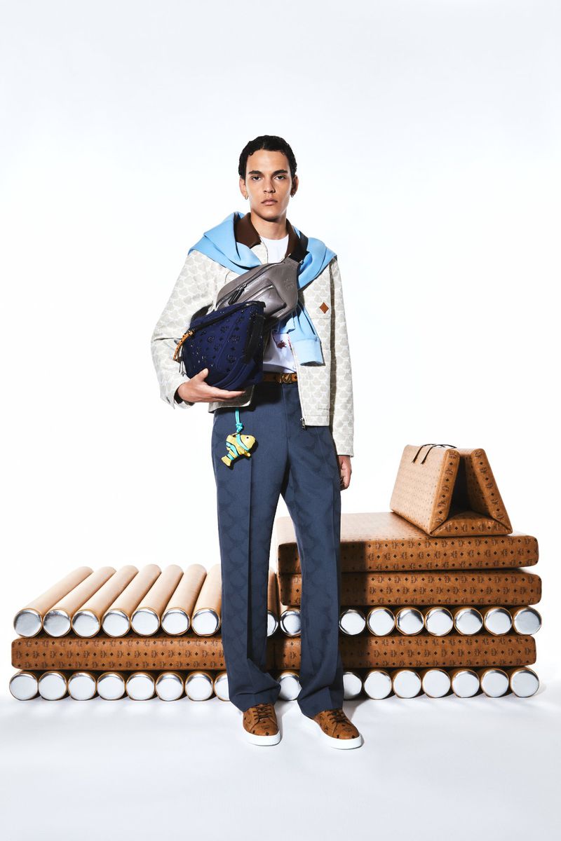 Full-length studio shot of a male model showcasing a designer outfit, including jackets, pants, shoes, and crossbody bag, on a white backdrop.