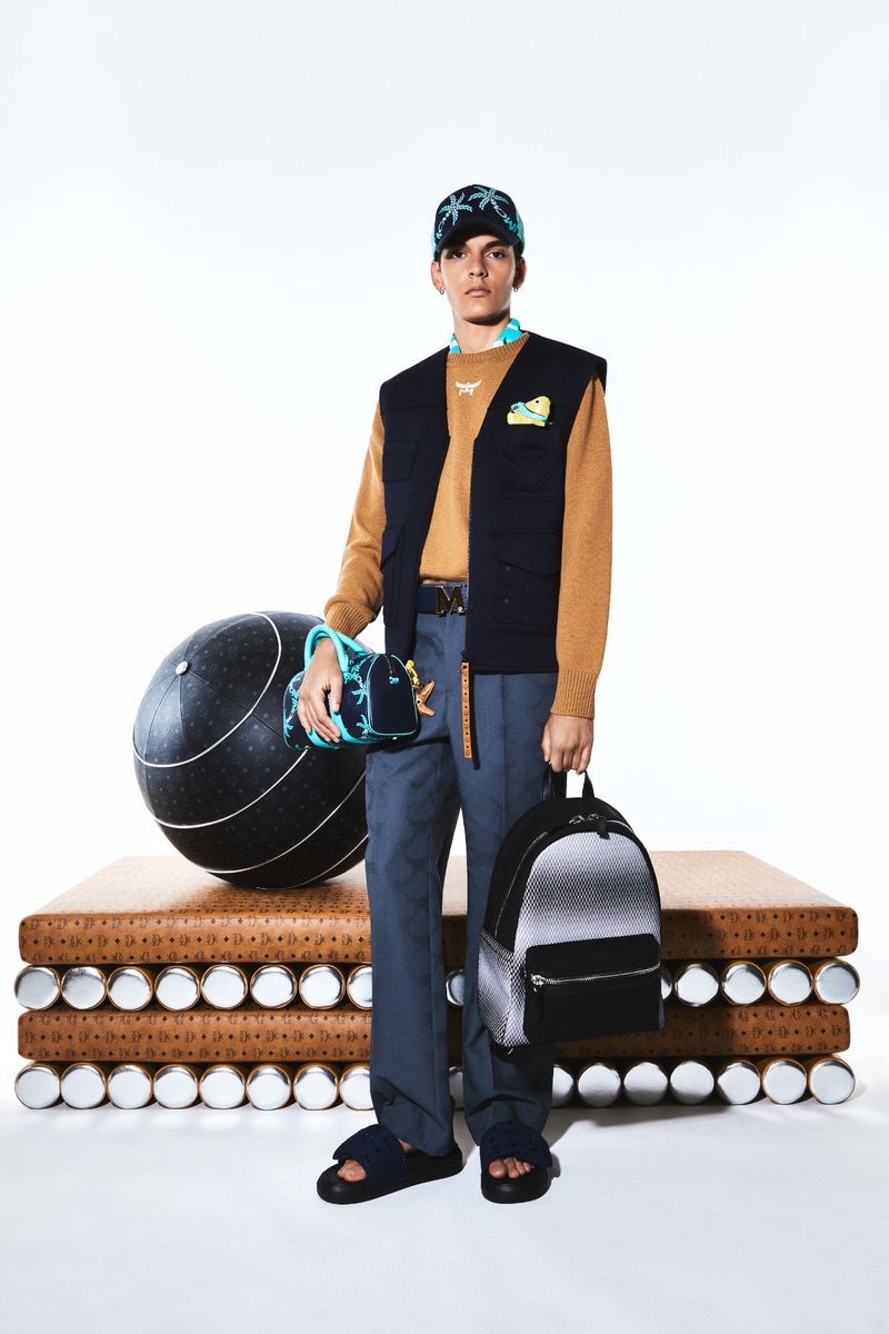 A young adult male model poses in a studio wearing a vest, sweater, and pants with luxury brand accessories, including a backpack and ball.