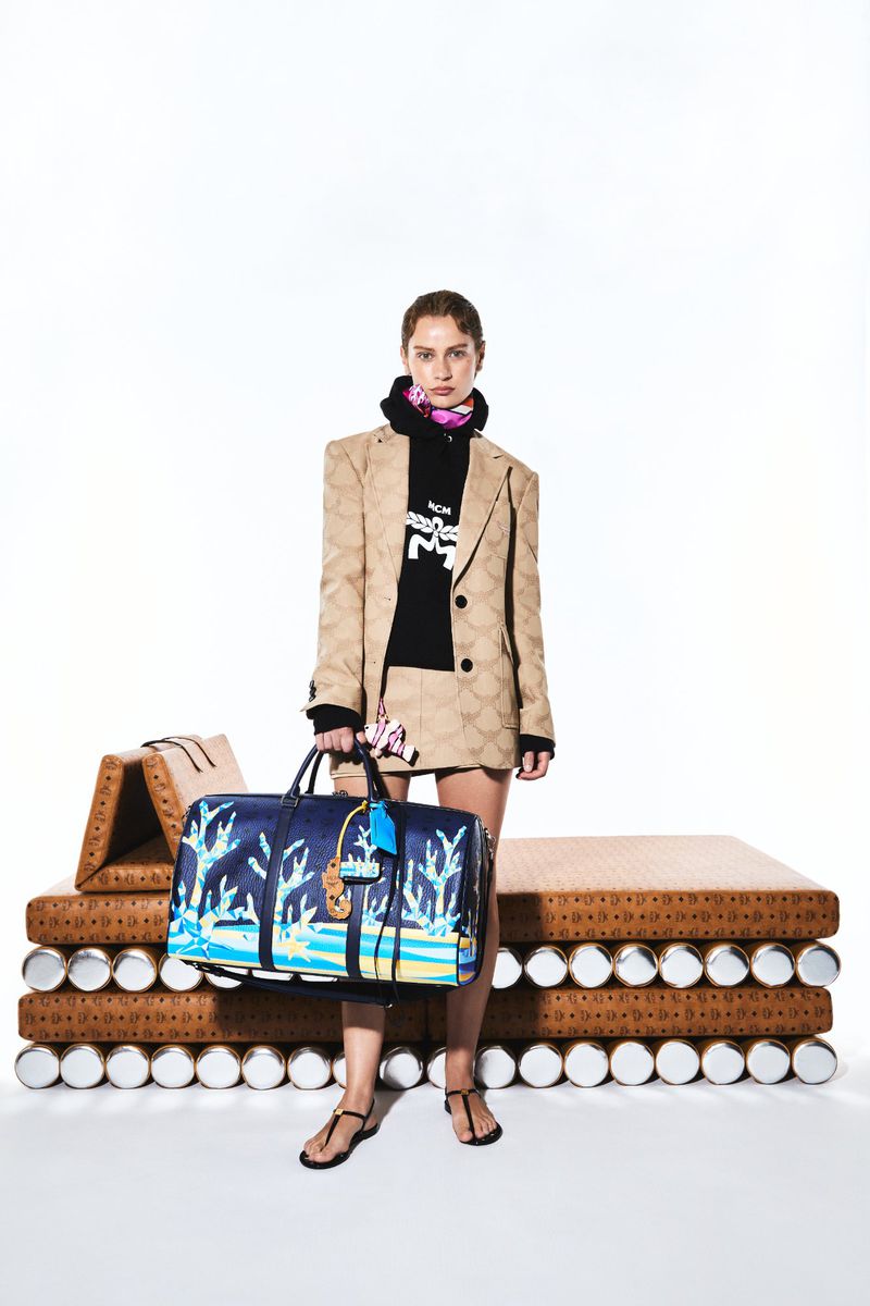 A woman in a patterned suit stands in a bright studio, holding a blue duffel bag with graphics. A platform is behind her.