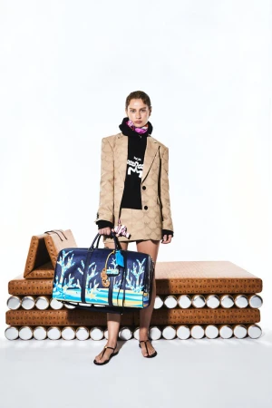 A woman in a patterned suit stands in a bright studio, holding a blue duffel bag with graphics. A platform is behind her.
