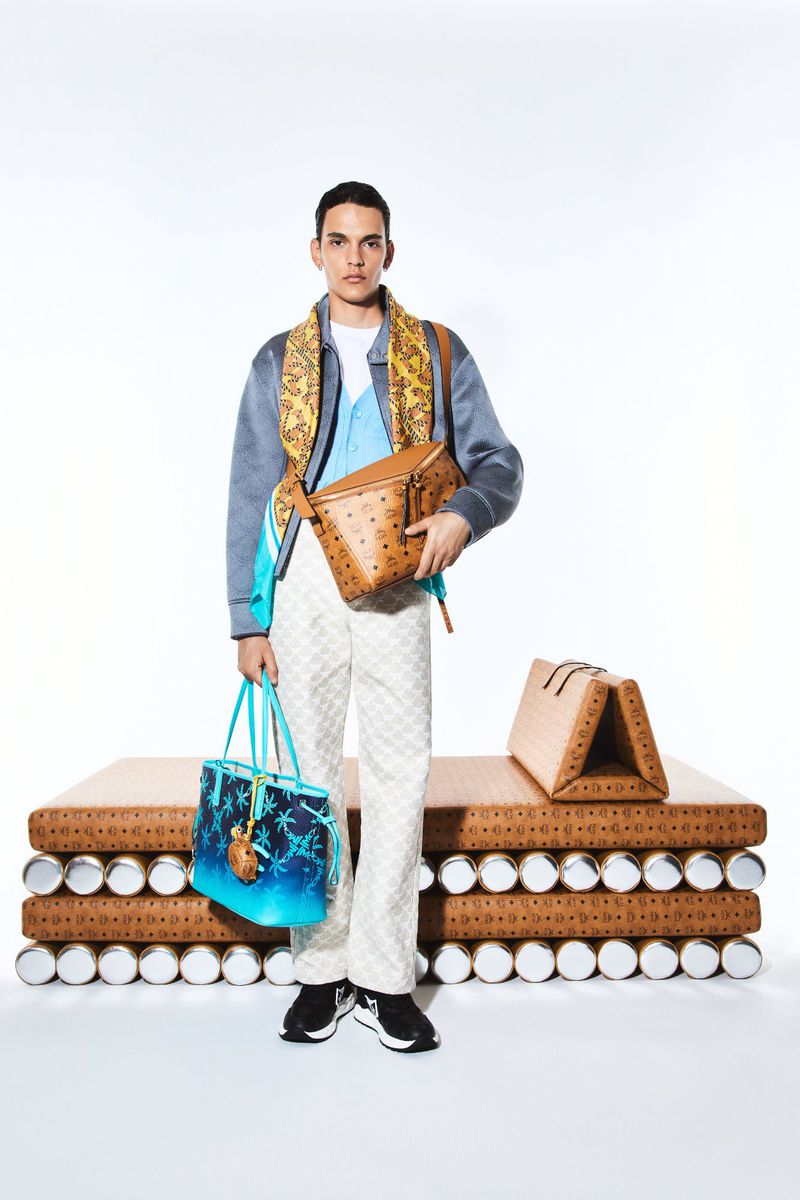 A fashionable adult male poses in a studio, wearing a patterned jacket, scarf, patterned pants, black sneakers, and carrying multiple handbags.