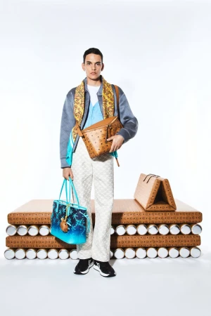 A fashionable adult male poses in a studio, wearing a patterned jacket, scarf, patterned pants, black sneakers, and carrying multiple handbags.