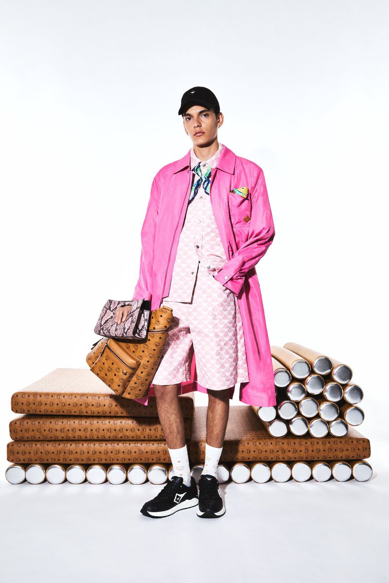 A young adult male fashion model strikes a pose in a pink coat and patterned shorts, accessorized with MCM bags and a baseball cap.