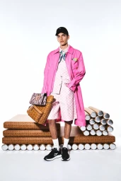 Male model in pink jacket and patterned shorts holding a monogram backpack against a white backdrop