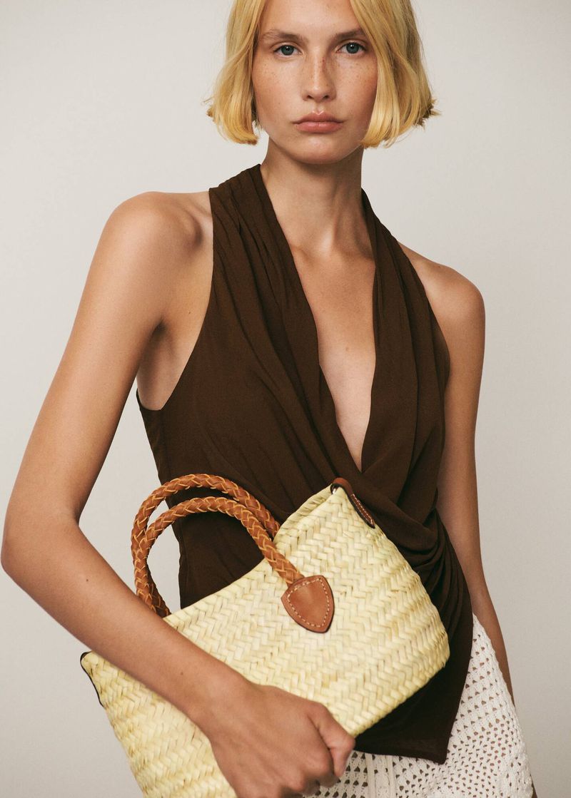 A fair-skinned woman with a brown halter top holds a straw tote bag in a studio setting against a neutral-colored background.