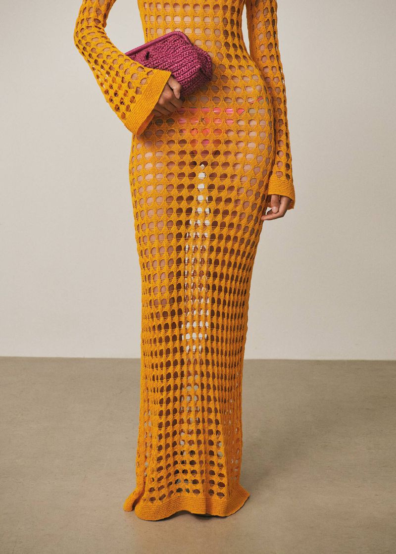 Adult woman wearing a mustard-colored crocheted dress and holding a small purple knitted handbag, standing in a studio with a neutral backdrop.