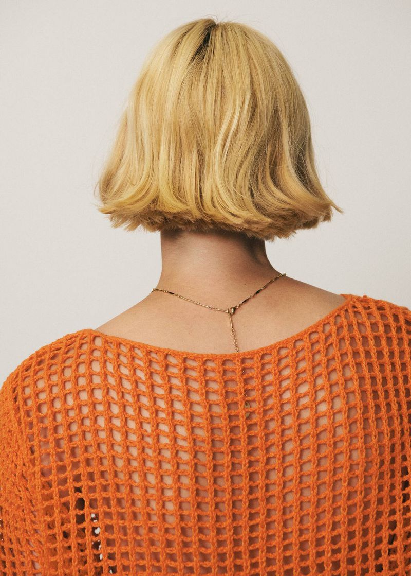 Back view of a woman with short blonde hair wearing an orange, textured knit top and gold necklace against a light grey background.