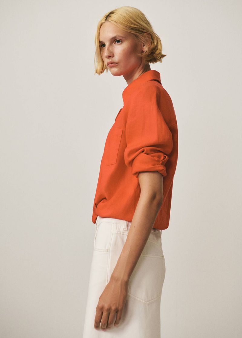 A slender woman with short blonde hair poses indoors wearing an orange button-down shirt and white skirt against a neutral backdrop.