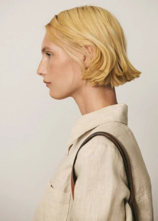 Profile shot of a fair-skinned adult female with short blonde hair, wearing a beige linen shirt and brown leather purse strap indoors.