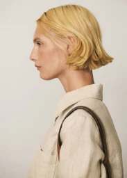 Profile portrait of a blonde woman wearing a light beige shirt and carrying a brown leather bag against a neutral background.