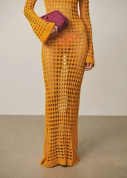 Model wearing a yellow open-knit maxi dress and holding a pink woven clutch bag against a plain studio background.