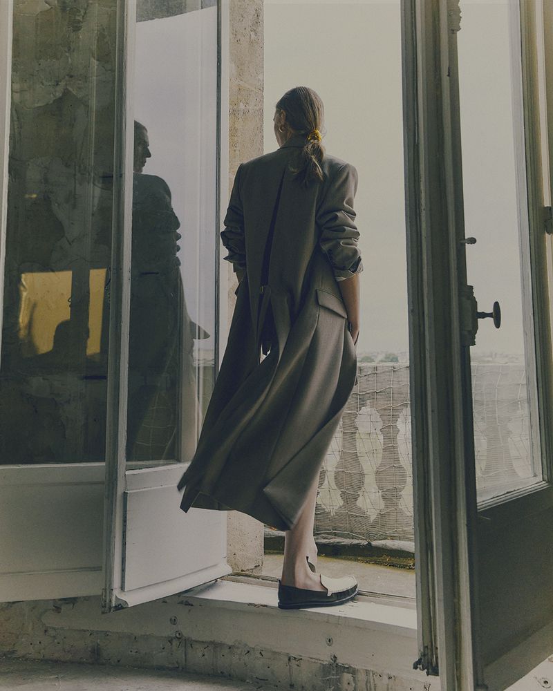 Woman in long grey coat stands in doorway overlooking a city skyline on a cloudy day, with reflection in a glass door.