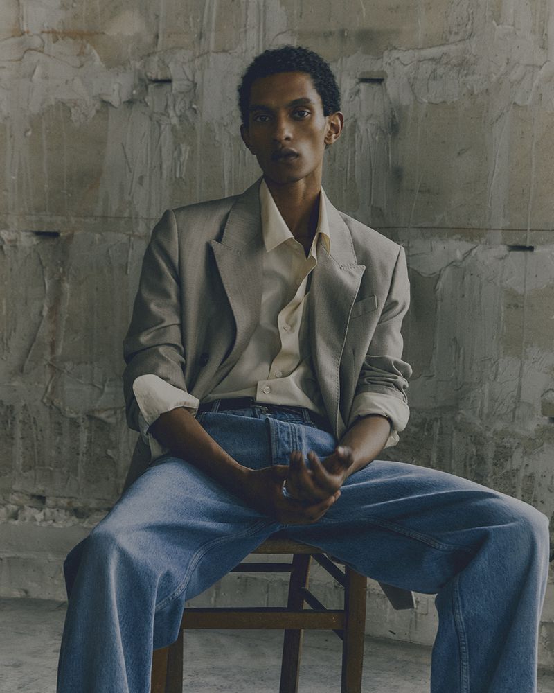Man in a blazer and jeans sits on a wooden chair against a textured concrete wall in a portrait-style fashion shot.