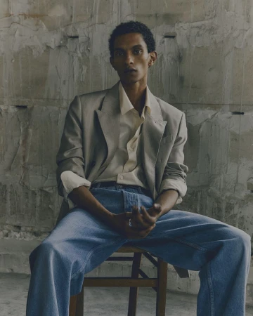 Man in a blazer and jeans sits on a wooden chair against a textured concrete wall in a portrait-style fashion shot.