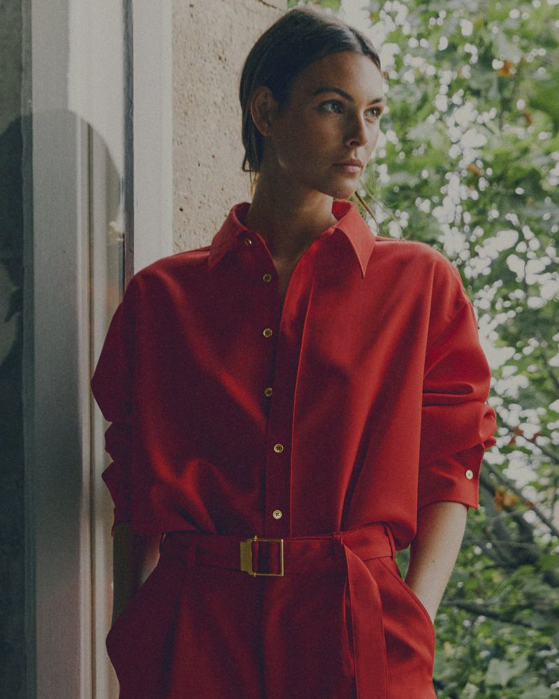 A woman models a vibrant red shirt and matching pants with a gold buckle belt, standing indoors near a window and greenery.