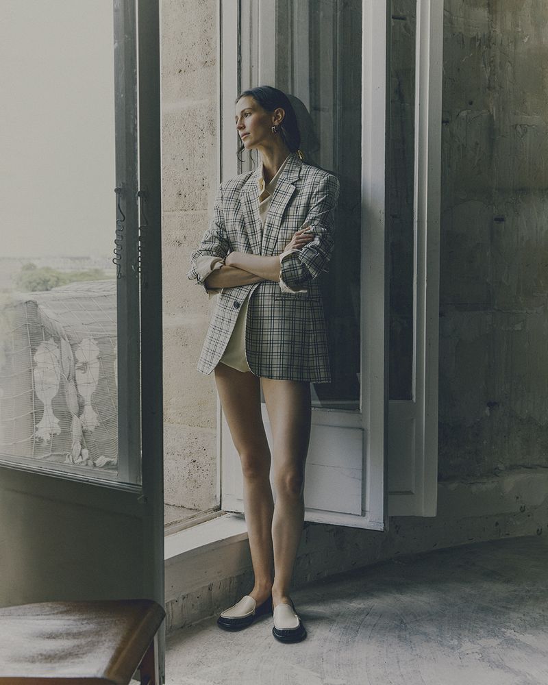 A stylish, slender woman in a blazer poses in front of an open window with stone walls and muted colors, creating an editorial feel.