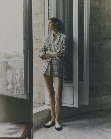 A stylish, slender woman in a blazer poses in front of an open window with stone walls and muted colors, creating an editorial feel.