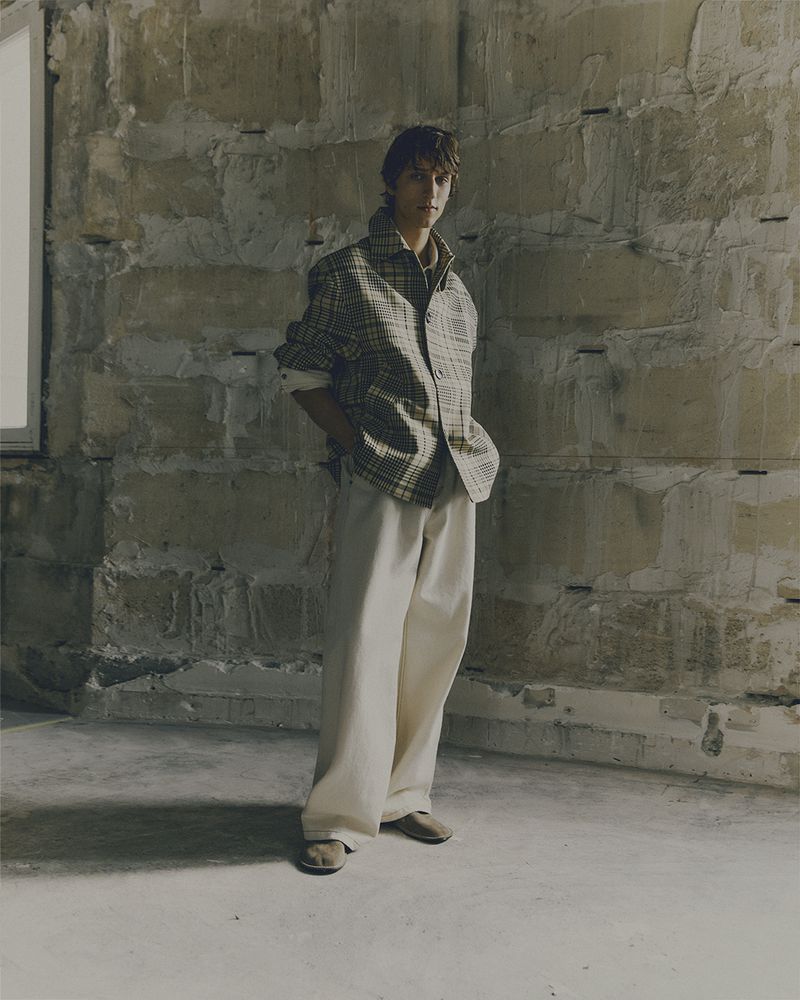 Full-length portrait of a young adult man in plaid shirt and wide-leg pants standing in an empty stone-walled room in neutral lighting.