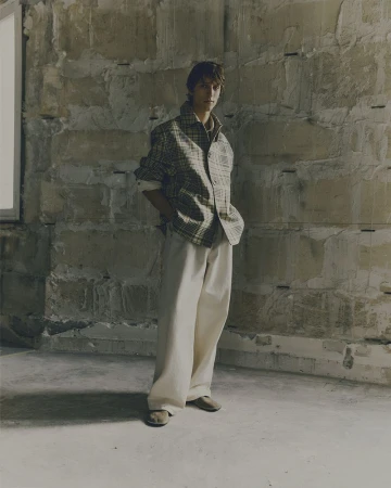 Full-length portrait of a young adult man in plaid shirt and wide-leg pants standing in an empty stone-walled room in neutral lighting.