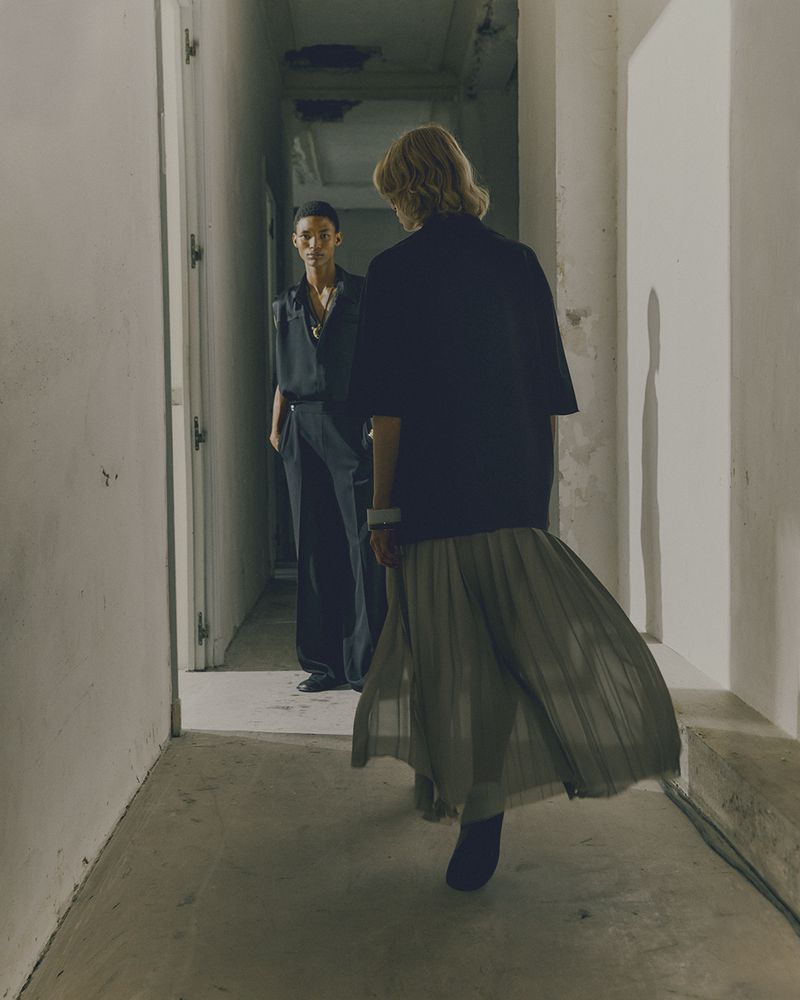 Two fashion models in a dimly lit hallway. One woman faces the camera wearing a long pleated skirt. The other looks at her.