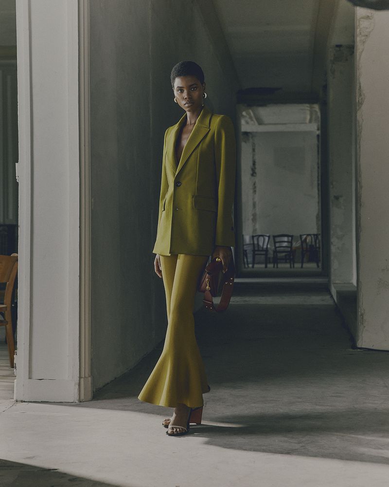 Woman in olive blazer and flared pants poses in corridor. Fashion shot. Gold hoop earrings. Neutral color palette. Shadows.