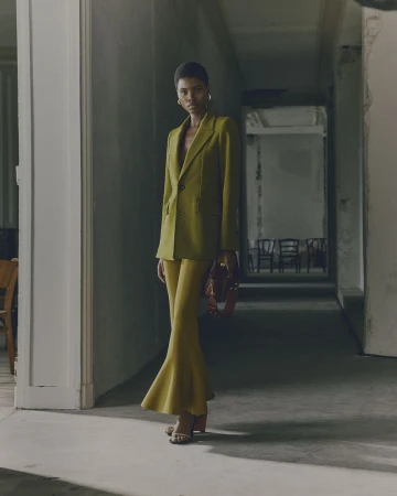 Woman in olive blazer and flared pants poses in corridor. Fashion shot. Gold hoop earrings. Neutral color palette. Shadows.