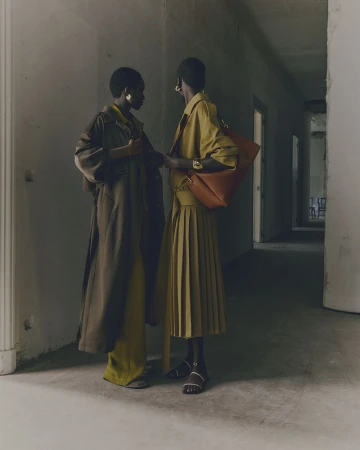 Two Black women in stylish coats and dresses converse in a minimalist hallway setting. One carries a matching handbag, creating a fashion-forward scene.