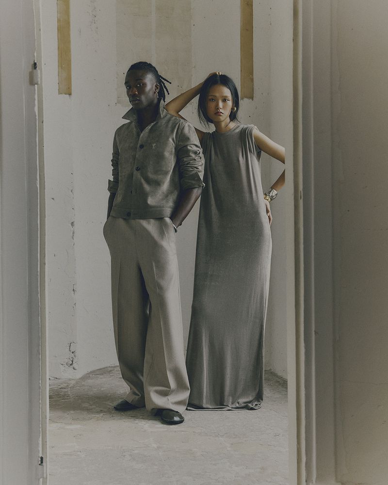 A Black man and an Asian woman pose in a dimly lit hallway, wearing complementary grey casual wear. The scene evokes an editorial aesthetic.