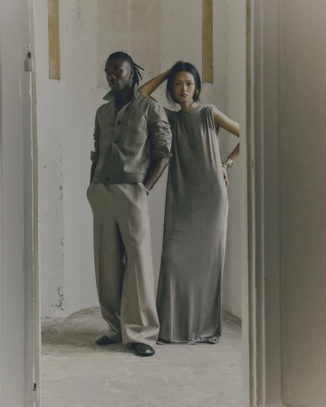 A Black man and an Asian woman pose in a dimly lit hallway, wearing complementary grey casual wear. The scene evokes an editorial aesthetic.