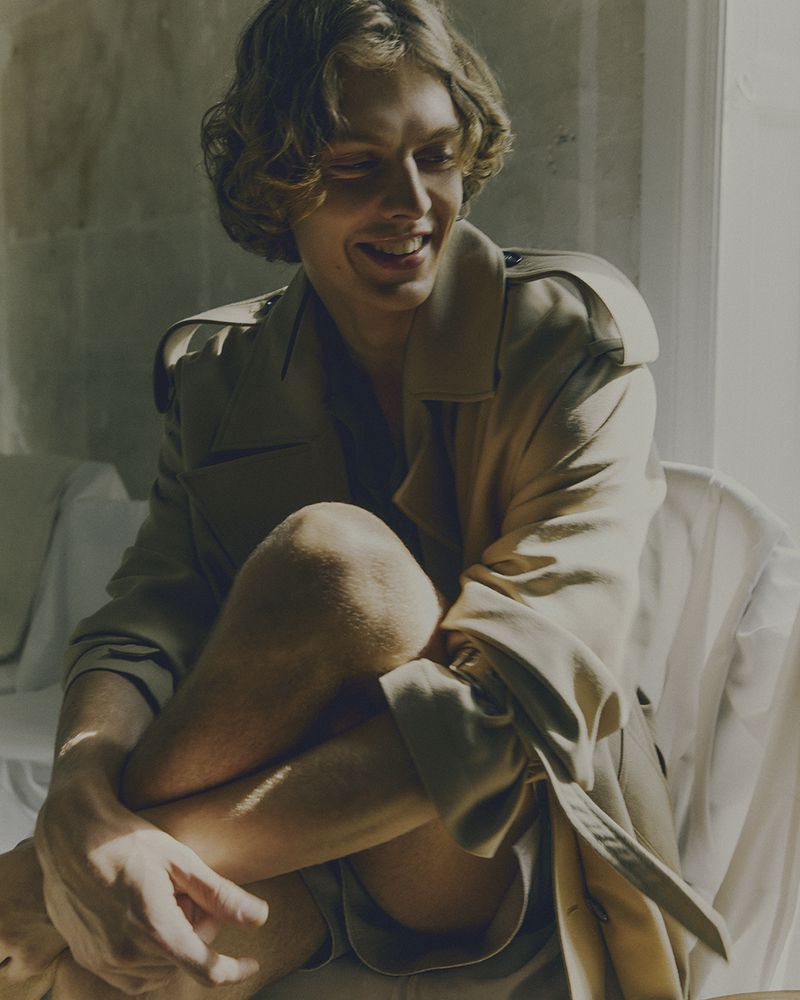Smiling man with curly hair poses indoors wearing a trench coat. He's sitting with arms wrapped around his legs in natural light.