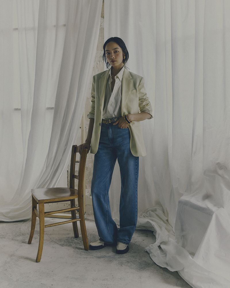 Full-length shot of a stylish woman in a blazer and jeans standing next to a chair in front of draped sheer white fabric.