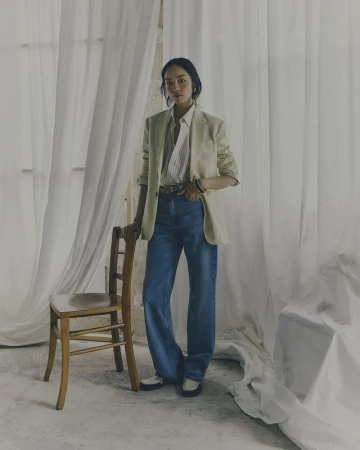 Full-length shot of a stylish woman in a blazer and jeans standing next to a chair in front of draped sheer white fabric.