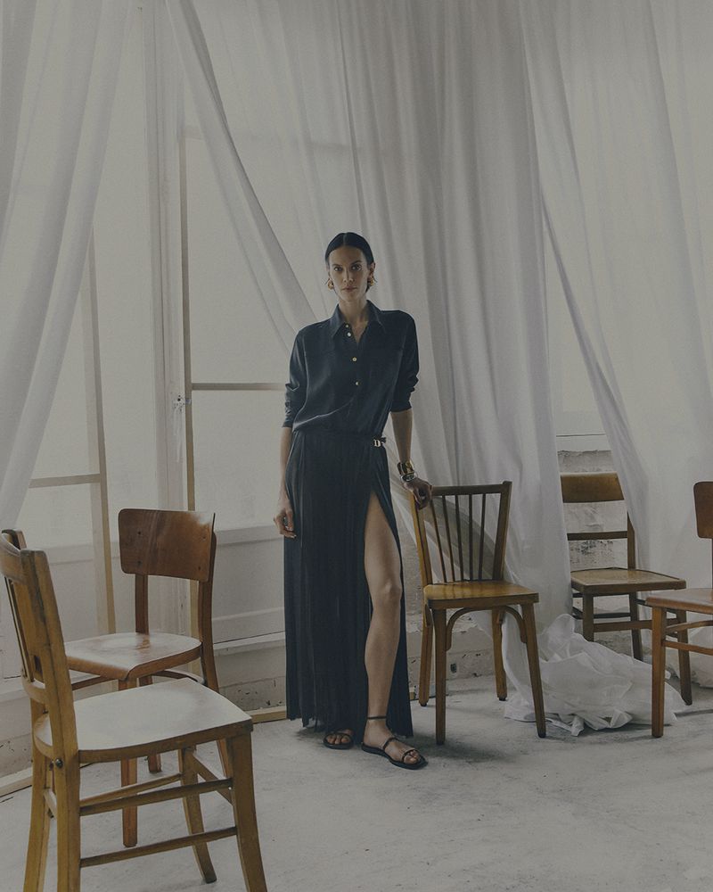 A woman in a dark shirt and long skirt stands amid an arrangement of wooden chairs in a bright, open space with sheer curtains.