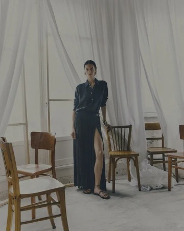 A woman in a dark shirt and long skirt stands amid an arrangement of wooden chairs in a bright, open space with sheer curtains.