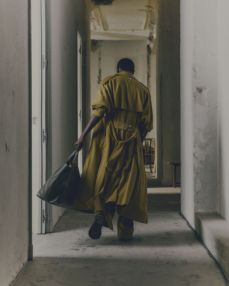 An adult strides down a hallway in a yellow trench coat, carrying a black bag, captured with neutral lighting and architectural backdrop.