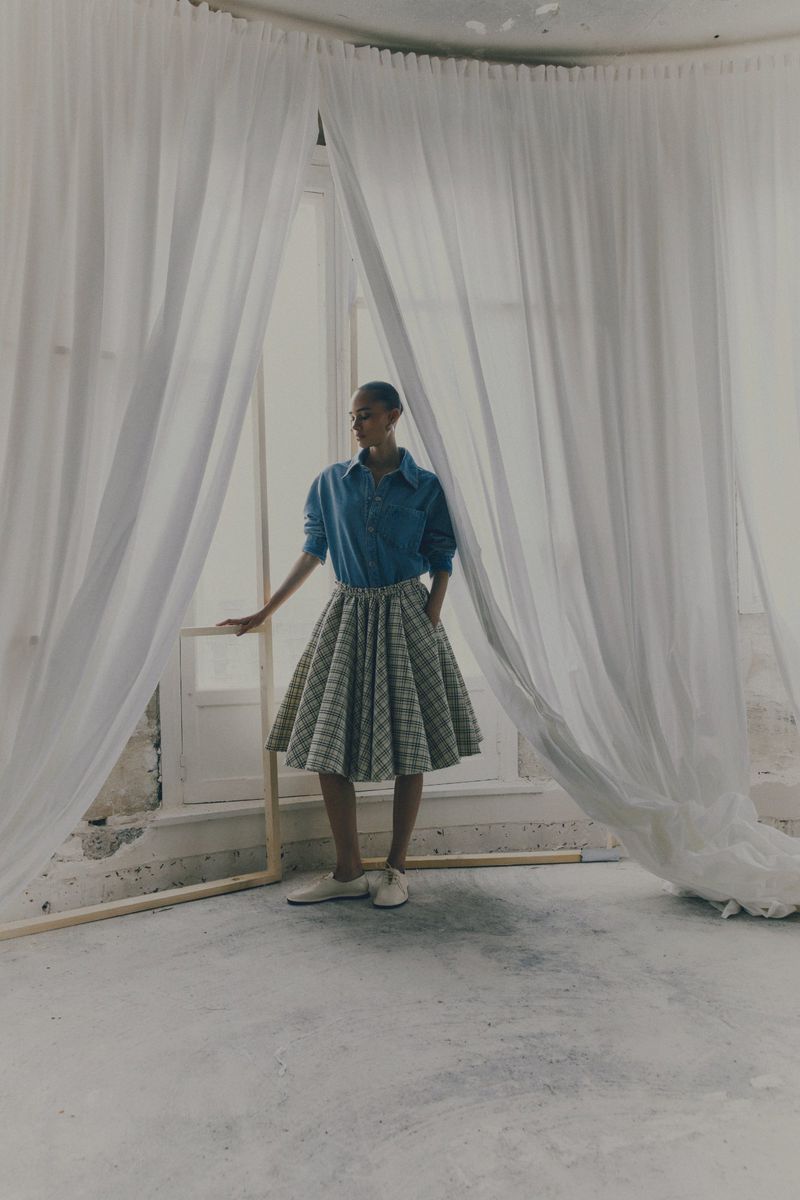 Woman in blue button-down shirt and pleated skirt standing by a window draped with sheer white curtains in a minimalist room.