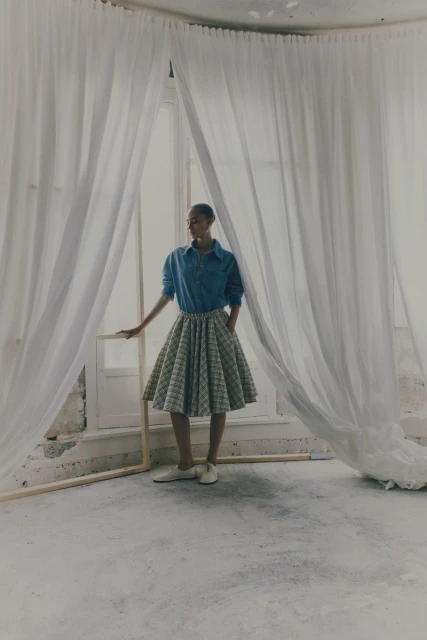 Woman in blue button-down shirt and pleated skirt standing by a window draped with sheer white curtains in a minimalist room.