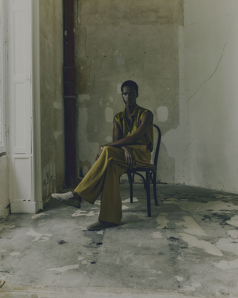 A person sits on a black chair in a sparsely decorated room, wearing mustard-colored clothing, with a stone floor and peeling paint.
