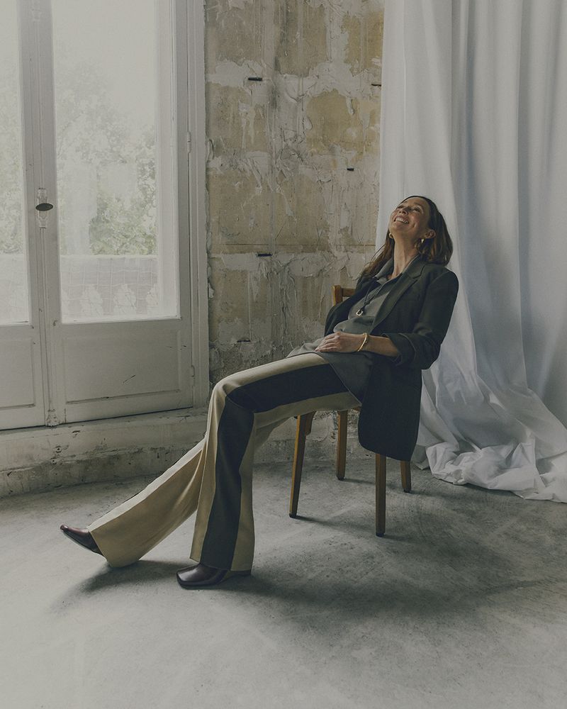 A woman in a blazer and color-blocked pants sits on a wooden chair, smiling in a bright, naturally-lit room with a rustic wall.